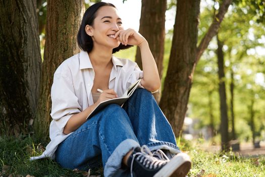 Portrait of young asian woman writing in her notebook, expressing her thoughts on paper in diary, smiling and sitting under tree on summer day in park.