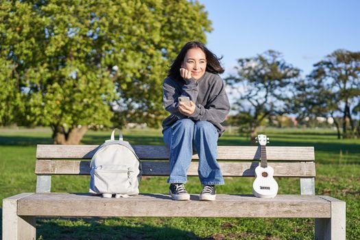 Young woman with ukulele, sitting on bench in park, using mobile phone app, holding smartphone and smiling.