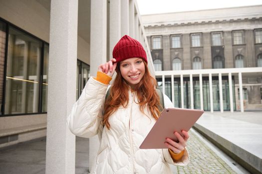 Stylish ginger girl, tourist walks with digital tablet around city, woman connects to iternet on her gadget, looking up information, texting message.