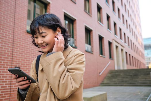 Smiling brunette woman, asian student walks on street and listens music in wireless earphones, holds smartphone, reads text message on mobile phone, picks song from playlist.