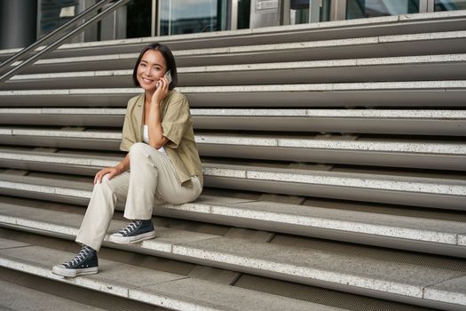 Smiling asian girl sits on stairs of building and talks on mobile phone, relaxing during telephone conversation.