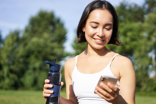 Smiling fitness girl drinks water, checks her app on smartphone and looks happy, stays hydrated on fresh air, sunny day in park.