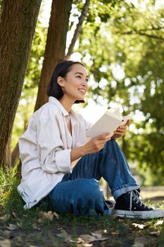 Vertical shot of happy asian woman relaxing outdoors in park, reading her book and sitting under tree shade on warm sunny day.