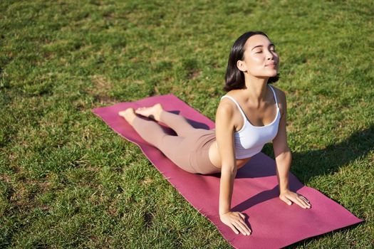 Smiling young sportswoman stretches on rubber mat in park, does yoga asana exercises, workout on fresh air in fitness clothing.