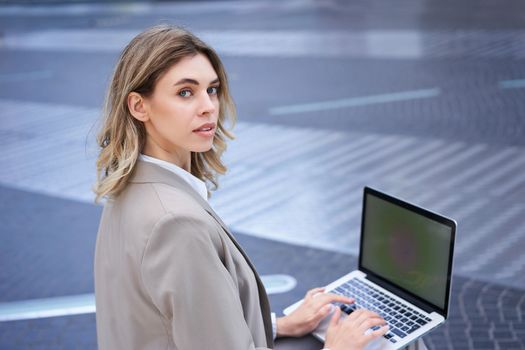 Young businesswoman using laptop while sitting outdoors in city centre, typing on keyboard. Girl preparing for interview, wearing suit.