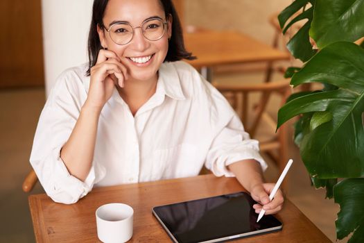 Young asian creative girl, sitting with digital tablet, drawing with graphic pen and smiling, doodling, drinking coffee in cafe.