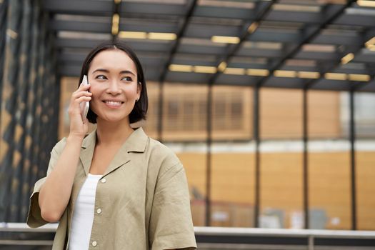 Happy asian woman talking on mobile phone, standing on bus stop in city. Outdoor shot.