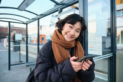 Cute smiling asian girl standing on bus stop, holding smartphone, wearing winter jacket and scarf. Woman commuting to work or university via public transport, stands on road.