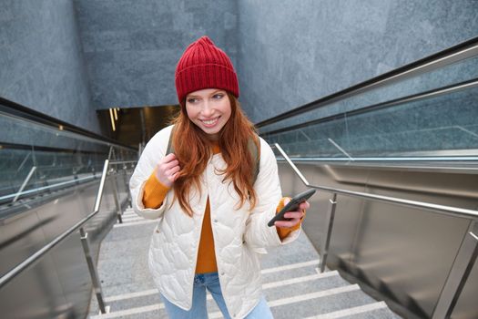 Outdoor shot of young woman plans route, follows map on smartphone app, goes up stairs with backpack and smiles.