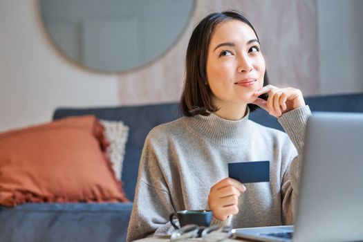 Happy smiling girl with credit card, paying her bills online on computer, doing shopping on her laptop, sitting at home.