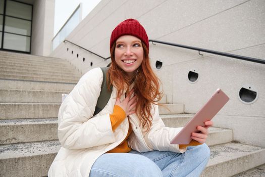 Young stylish girl, redhead female students sits on stairs outdoors with digital tablet, reads, uses social media app on gadget, plays games while waits on street.