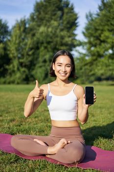 Vertical shot of asian woman shows thumbs up, recommending yoga training online, meditation app, doing exercises on fresh air in park, showing smartphone screen.