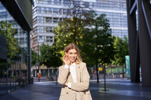 Portrait of smiling businesswoman in corporate clothing, looking confident and happy, wearing beige suit, standing outdoors on street, outside office.