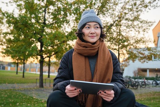 Smiling young girl, korean student sits in park in scarf and hat on chilly day, draws outdoors on digital tablet with graphic pen, makes scatches.