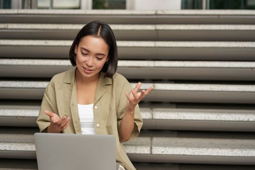 Portrait of young asian girl, student talks at laptop, video chat, speaking during online meeting, sitting outdoors on stairs.