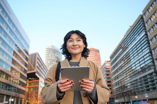 Portrait of happy korean girl smiles, walks in city with digital tablet and backpack, uses her gadget to get around.