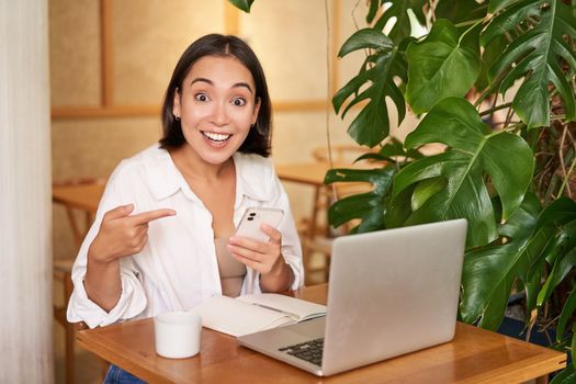 Amazed asian girl points at her phone and looks surprised, tells big news, sits in cafe with laptop.