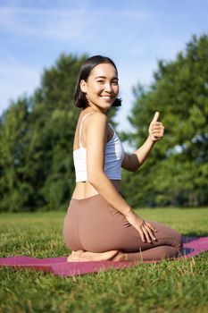 Portrait of asian woman say yes to yoga training in park, makes thumb up, sitting and meditating.
