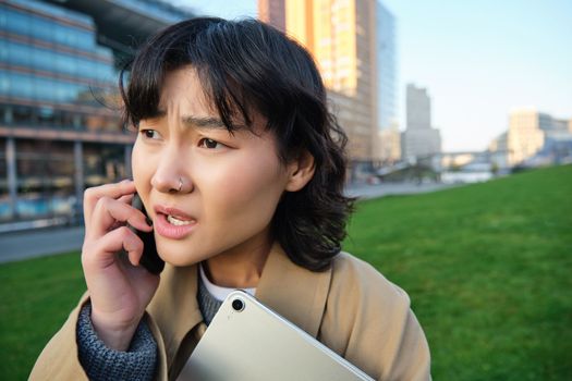 Close up portrait of young asian woman talks on mobile phone with concerned face, hear bad news over telephone conversation, walks from university with tablet.