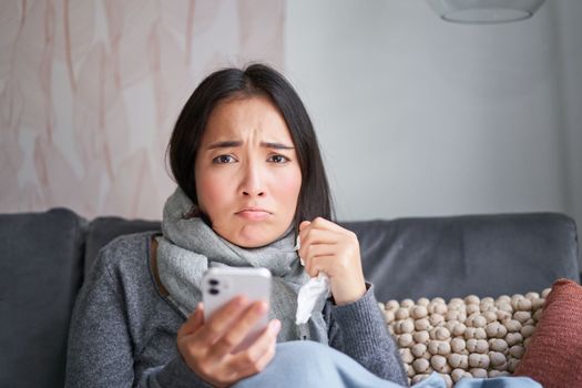 Asian girl with influenza, sits at home, cries and looks upset, holds smartphone, feels unwell and gloomy, wearing warm clothes.