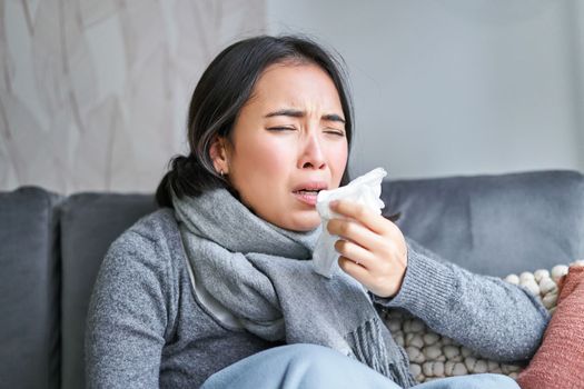 Close up of korean girl sneezing in napking, sitting at home in scarf, feeling unwell, concept of winter, heating cost and people.