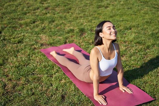 Smiling young sportswoman stretches on rubber mat in park, does yoga asana exercises, workout on fresh air in fitness clothing.