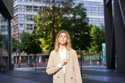 Portrait of young confident business woman in beige suit, corporate lawyer or saleswoman standing outside office building on street and smiling.