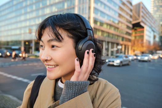 Portrait of young asian girl, student walks in city, listens music in headphones and uses mobile phone on streets.