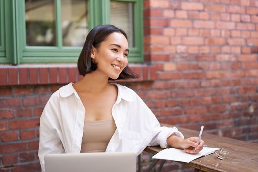 Beautiful young female model sitting with laptop in outdoor cafe, taking notes, writing in her diary or working on project, doing homework.