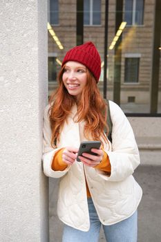 Stylish urban girl using mobile phone app, standing in city, waiting for taxi, looking at smartphone application, texting message.