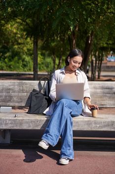 Portrait of asian woman sitting with laptop on bench in park, listening music with wireless headphones, doing homework, working on remote.