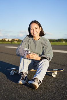 Vertical shot of asian woman sitting on skateboard on road, holding smartphone app. Skater girl skates on longboard, using mobile phone.