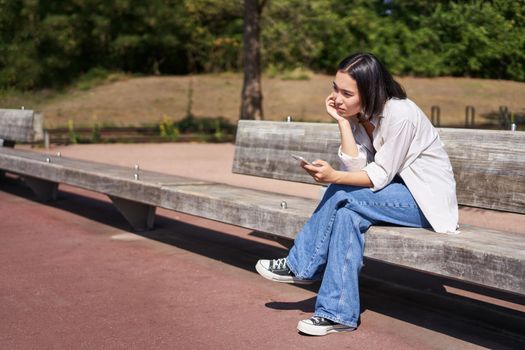 Portrait of asian girl sitting with smartphone feeling sad, looking gloomy and frustrated, waiting for a call outdoors in park.