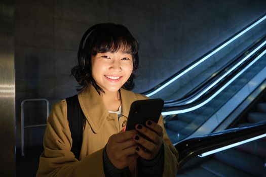 Stylish Japanese woman in headphones, standing with smartphone in hands near escalator, travelling and commuting in city, urban lifestyle concept.