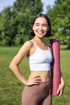 Smiling fitness girl with rubber mat, stands in park wearing uniform for workout and sport activities, does yoga outdoors on lawn.