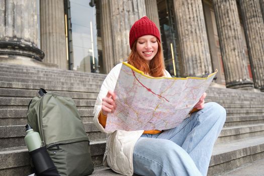 Portrait of young redhead woman, tourist sits with paper map and looks for a route to tourism attraction, rests on stairs outdoors.
