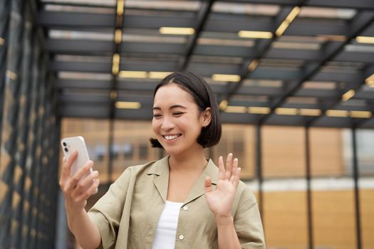 Happy asian girl waves at her smartphone camera, says hello to friend on video chat, calling someone, standing on street.