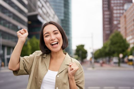 Happy asian girl winning and rejoicing, celebrating on streets of city, dancing from joy, triumphing and smiling.