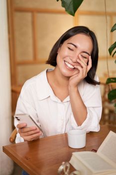 Stylish young woman in cafe with coffee, holding mobile phone, laughing and smiling. Lifestyle and people concept.