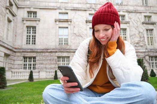 Portrait of stylish young woman, 25 years, sits on bench in park and uses mobile phone, reads online news, messages or watches video on smartphone app, connects to public wifi.