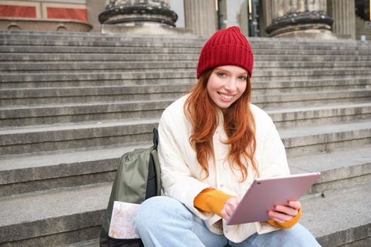 Beautiful young modern girl with red hair, holds digital tablet, sits on stairs near museum and connects public internet, sends message on gadget app.