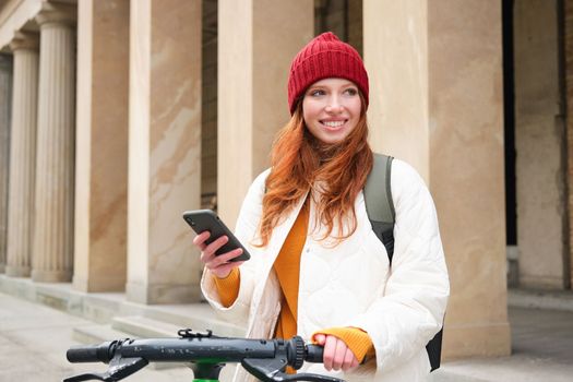 Redhead girl, tourist with backpack, uses mobile phone to rent e-scooter on streets of European city.