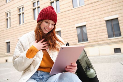 Redhead girl smiles, sits outdoors near building with digital tablet, thermos and backpack, connects to public internet and searches smth online on her gadget.