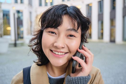 Portrait of smiling asian girl talks on mobile phone, speaks with friend on smartphone, walks in city centre, stands on street and laughs.