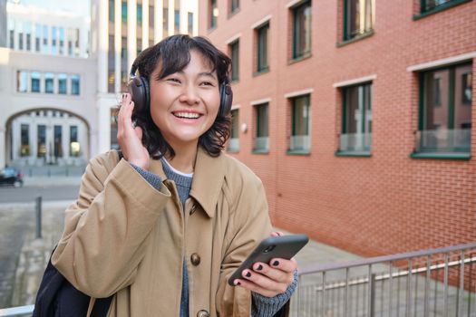 Young happy girl tourist, listens music in headphones, drinks takeaway and checks mobile phone, stands on street and smiles.