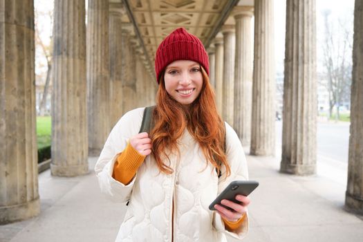 Happy young female model stands on street and holds mobile phone, uses smartphone app, follows the route of online map.