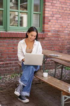 Vertical shot of asian woman working in outdoor cafe, sitting with laptop on bench and drinking coffee. Copy space