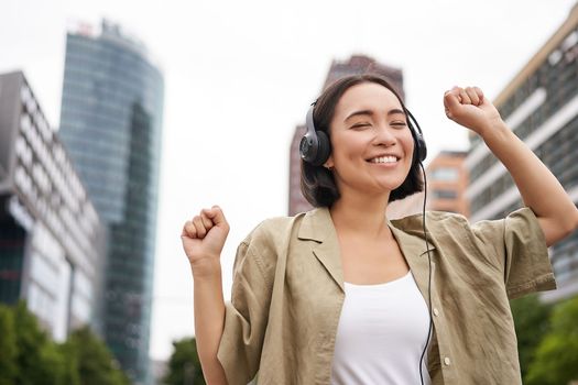 Happy asian woman in headphones, listening music and dancing on street of city centre, smiling with hands up.