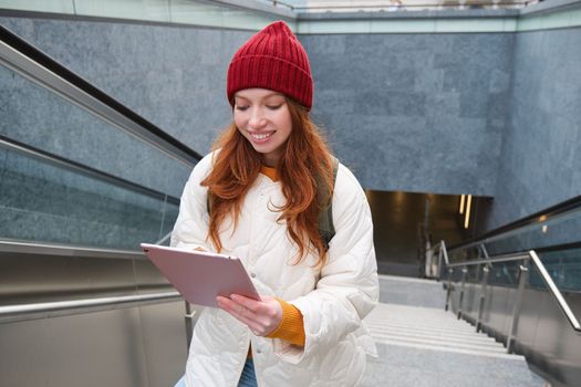 Smiling redhead woman walking up stairs with digital tablet, using gadget application while going somewhere in city.