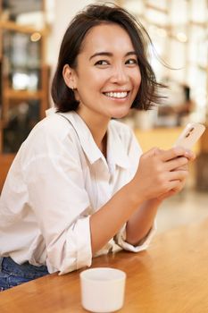 Vertical portrait of stylish asian woman sitting in cafe, drinking coffee and using smartphone. Lifestyle and people concept